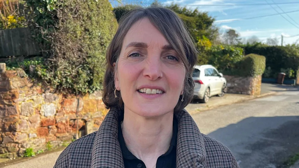 A woman with a nose ring and silver ear rings is standing in a sunny street in a town in Devon. She is wearing a brown checked coat with a black shirt underneath. 