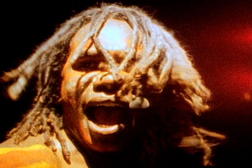 A young Aboriginal man with dreadlocked hair to the top of his shoulders turns his head. He wears body paint.