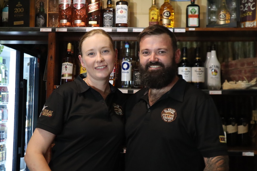 A woman and a man couple who own a pub standing in front of the bar with liqour bottles behind them.