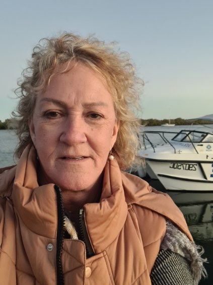 A caucasian woman wearing a brown vest jacket with a neutral expression stands in front of a boat
