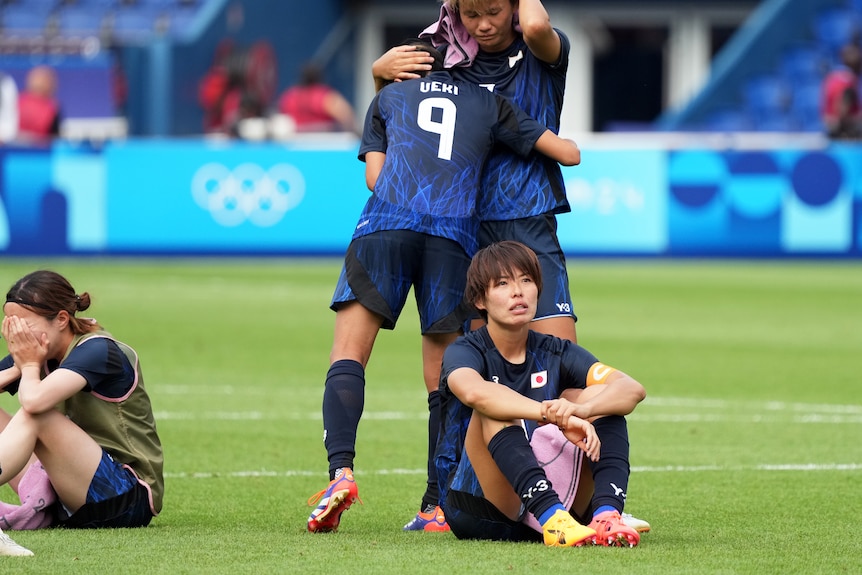 A player sits on the field and looks dejected as her teammates hug one another.