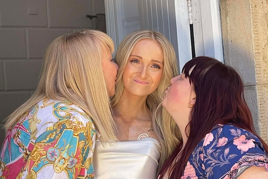A young woman in a wedding dress being kissed on the cheek by two women, one either side of her. 
