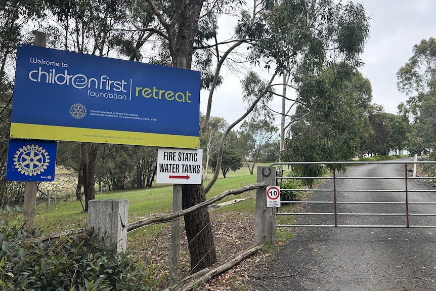 A sign next to a gate at the front of a driveway lined with trees.
