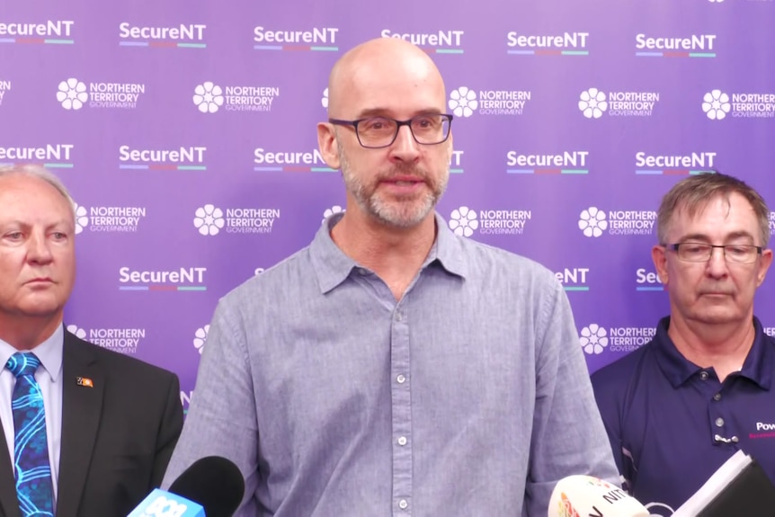 A white man in a blue button up shirt standing at a podium, purple banner behind him, two men behind him as well.