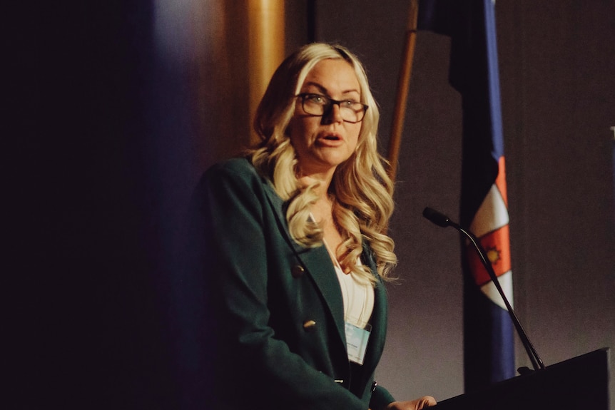 A woman in a suit stands talking at a lectern in front of flags