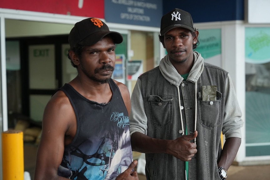 Two men wearing caps give thumbs up outside shops