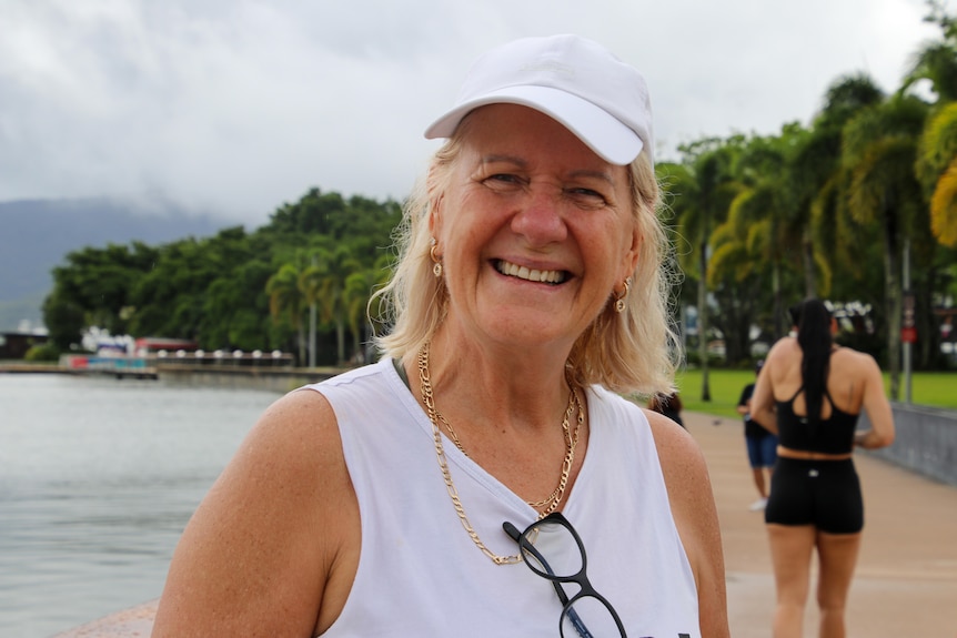 A smiling blonde woman in a cap and sleeveless top stands on a palm-lined esplanade.