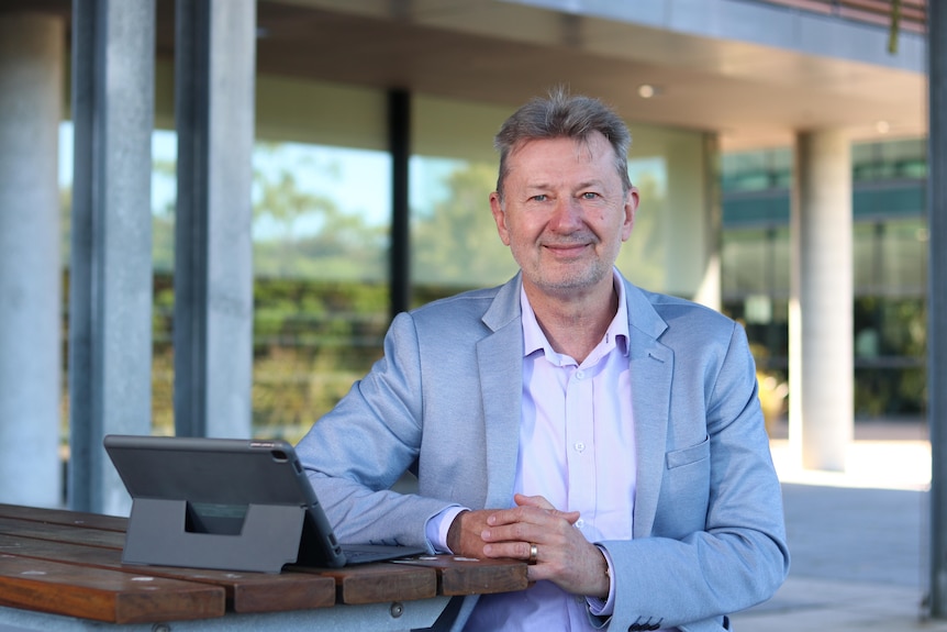 Smiling Ty, Caucasian, salt and pepper hair, in grey suit jacket and pink shirt, sits next to tablet, building behind.