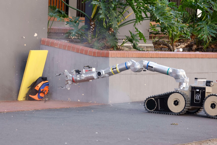 A robot on caterpillar wheels extends it arms towards a backpack on a footpath.
