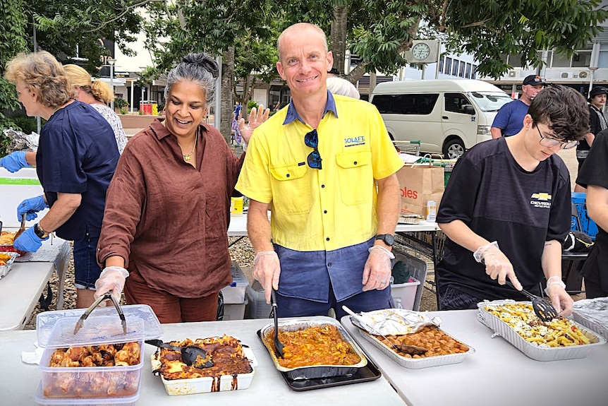 A woman in a maroon top and a man with a yellow shirt smile, and they stand behind a table with hot food.