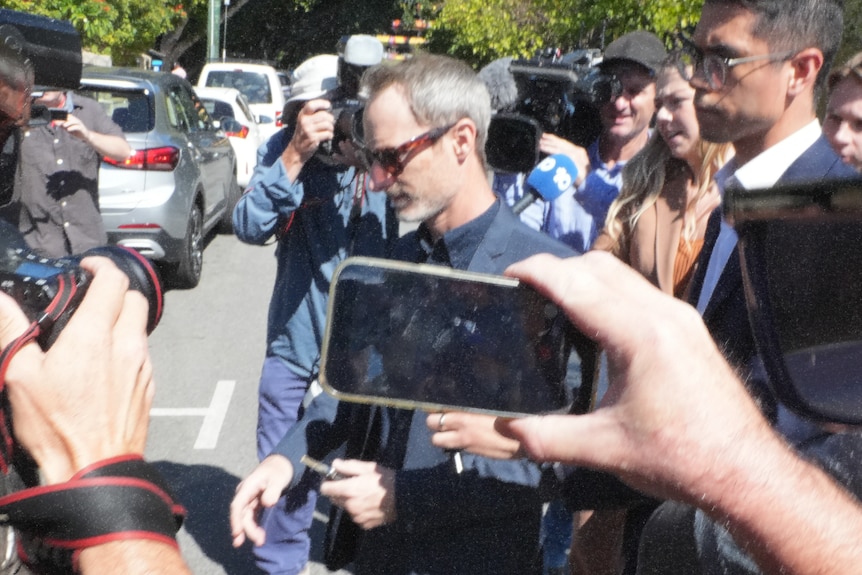 Craig Silvey crosses a road wearing sunglasses and a dark shirt surrounded by a media scrum and cameras.