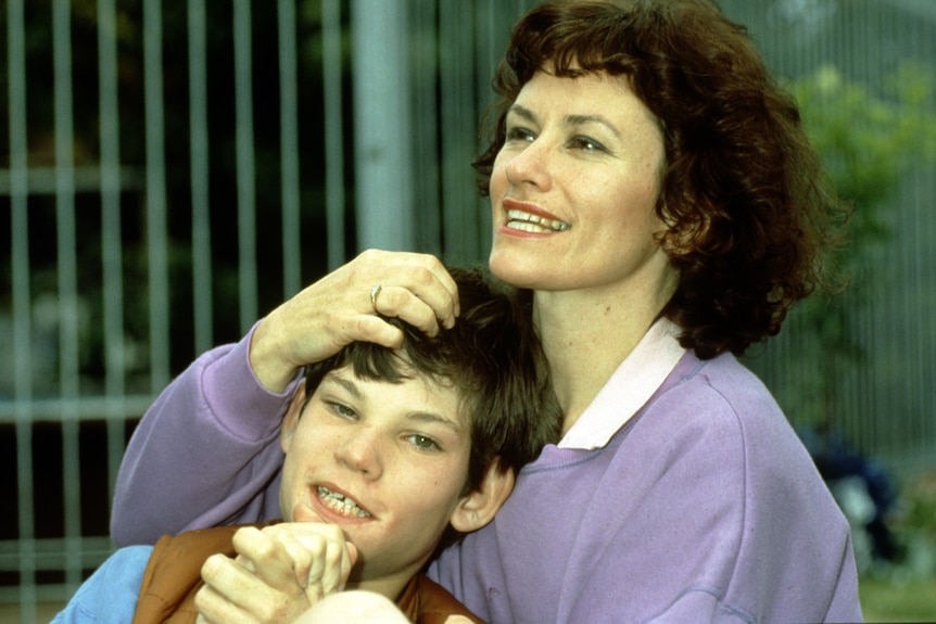 A young boy about age 10 clasps his hands together and smiles while his mum, brown curly hair, sits behind stroking his hair