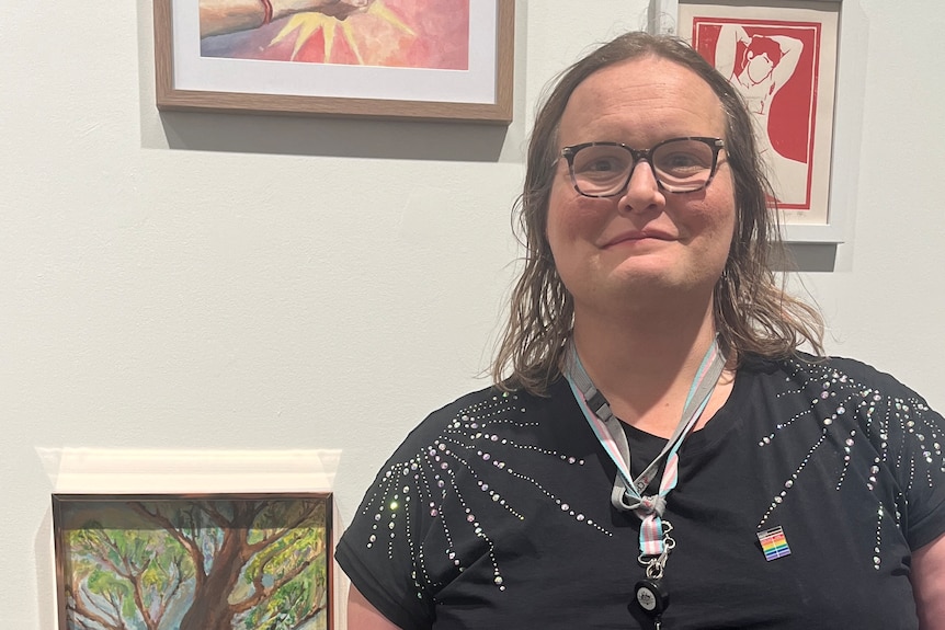 A dark-haired woman in glasses stands in front of a numb er of small paintings on a wall.