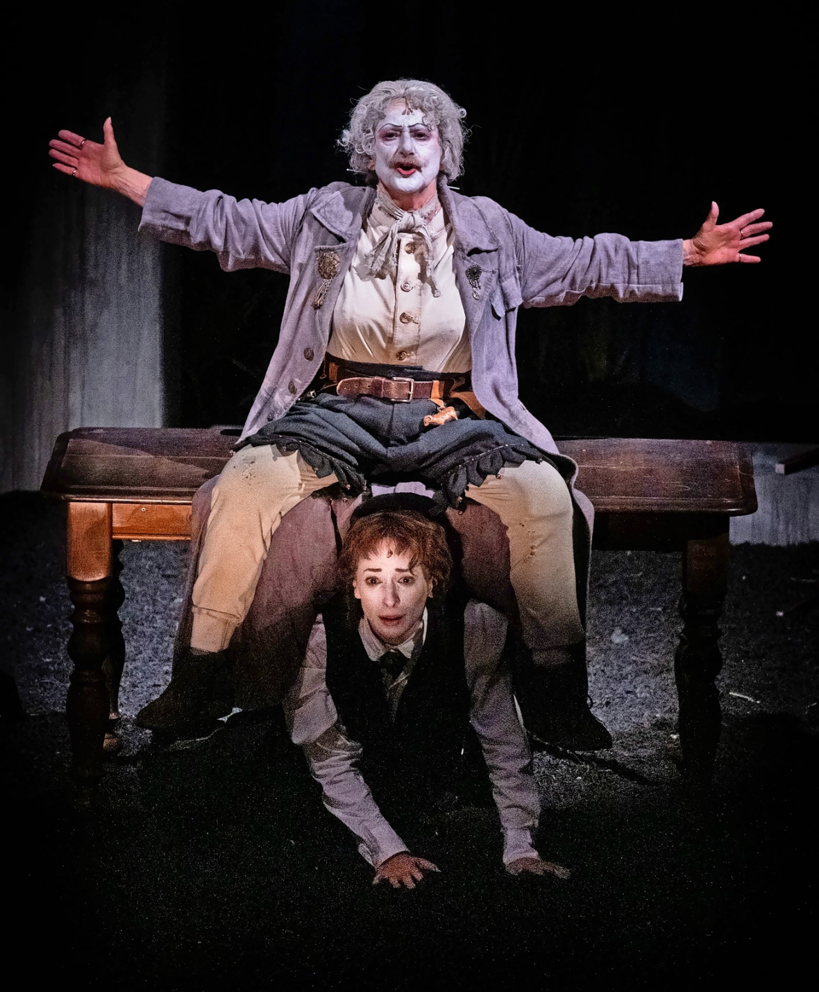 A person in clown makeup sits on a table, another person kneels below with a distressed expression.