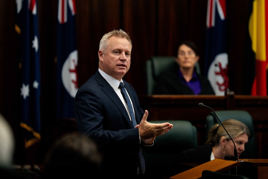 A man wearing a suit speaks with one hand outstretched, with two people and flags in the background.
