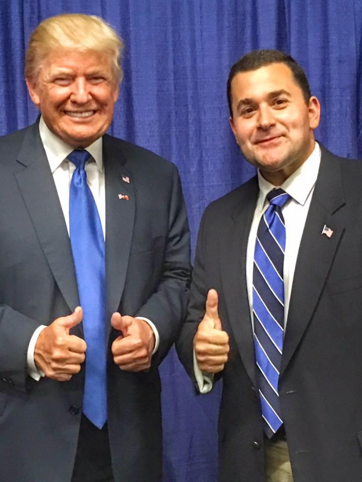 Donald Trump and Nick Adams wearing suits and smiling in front of a blue curtain