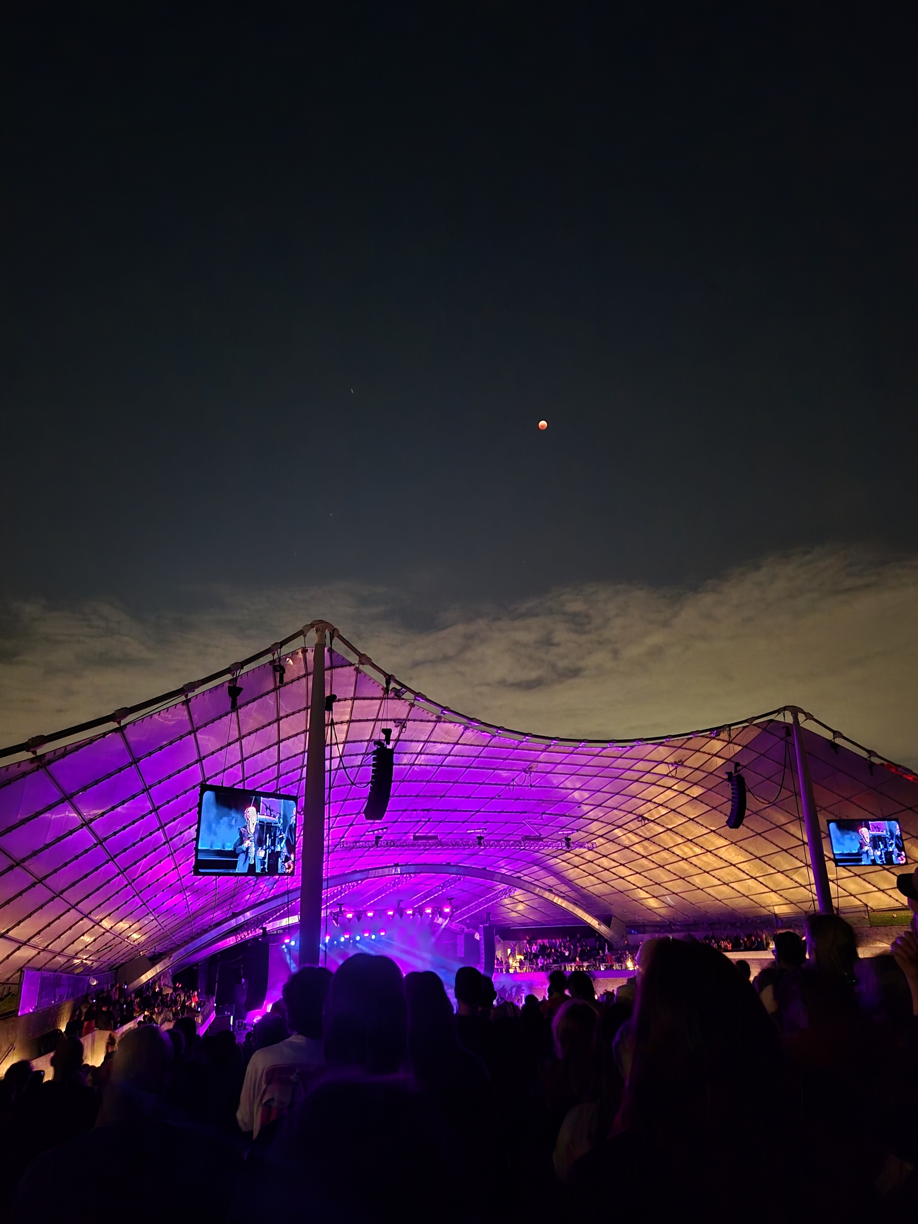 A bright orange eclipse moon high in the sky overlooking a purple tentage at an outdoor music performance.