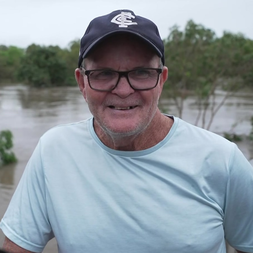 A man wearing glasses and a cap smiling, a swollen river can be seen in the background.