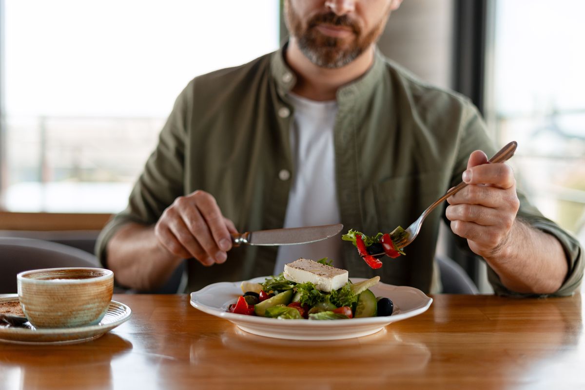 Mid adult man enjoying a fresh Greek salad topped with feta cheese while seated at a cozy restaurant table, savoring every bite