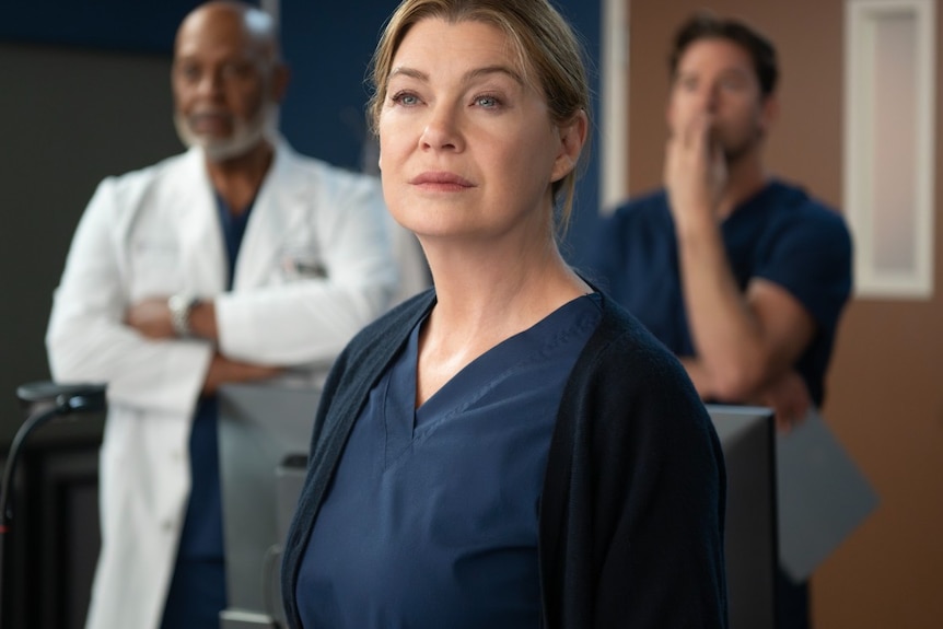 A woman in dark blue scrubs and a black cardigan, looking serious. She is flanked by two men in the background.