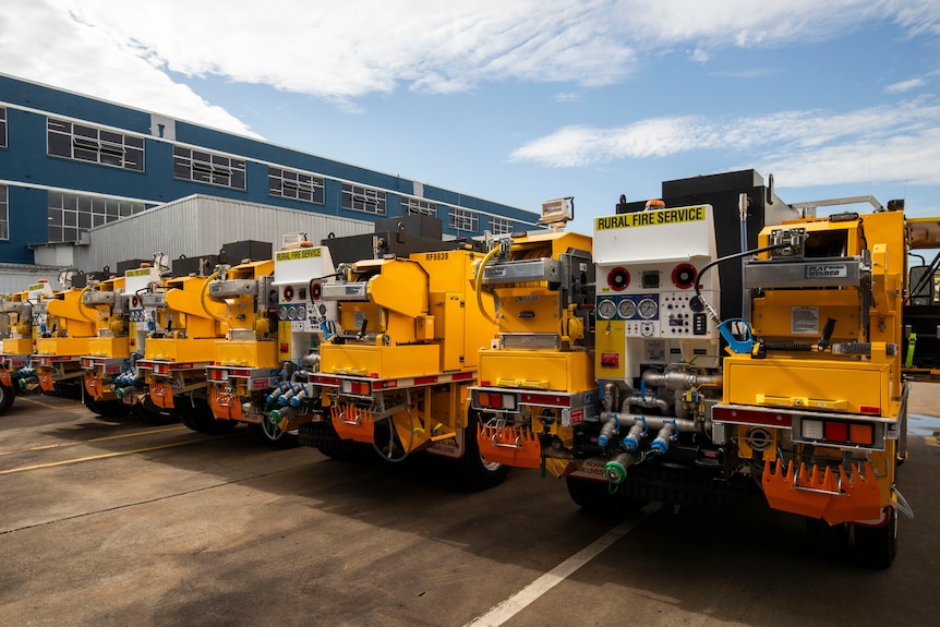 Medium-sized fire trucks are lined up in a carpark outside a two-storey fire service station. 
