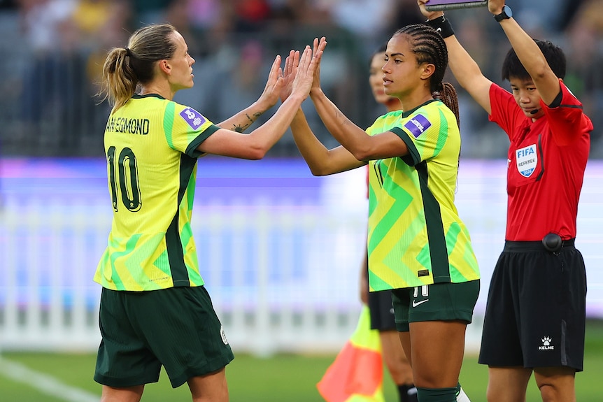 A Matildas player stands on the sideline and pats hands with her teammate who is coming off, as an electronic board is held up.