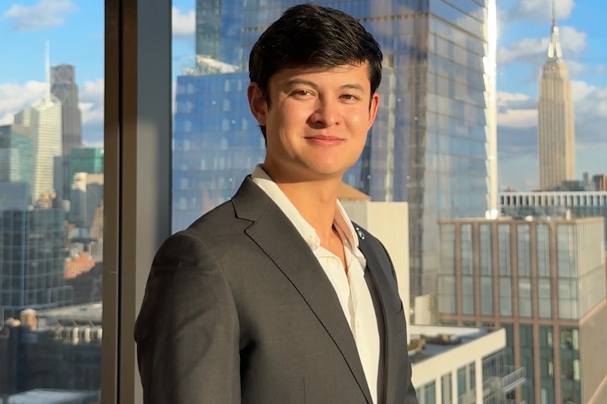 A man wearing a suit standing in an office with large windows and a cityscape behind him.