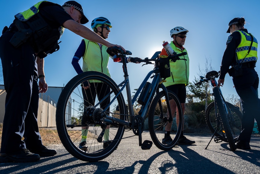 Two police officers inspect the bikes of two cyclists wearing helmets and hi-vi vest