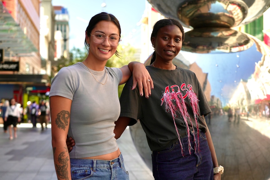 Two women stand together in a shopping mall. One rests her arm on the other's shoulder.