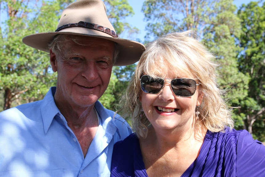 Mal Leyland age 69 wears a wide-brim hat, standing next to wide Laraine, blonde hair and sunglasses. Both smiling