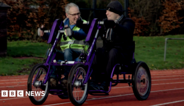 Two men, one on the left with grey hair and wearing glasses and another on the right wearing a hat, are sitting alongside each other on a type of bike, which is in part purple. It is on an athletics track and grass is in the background.