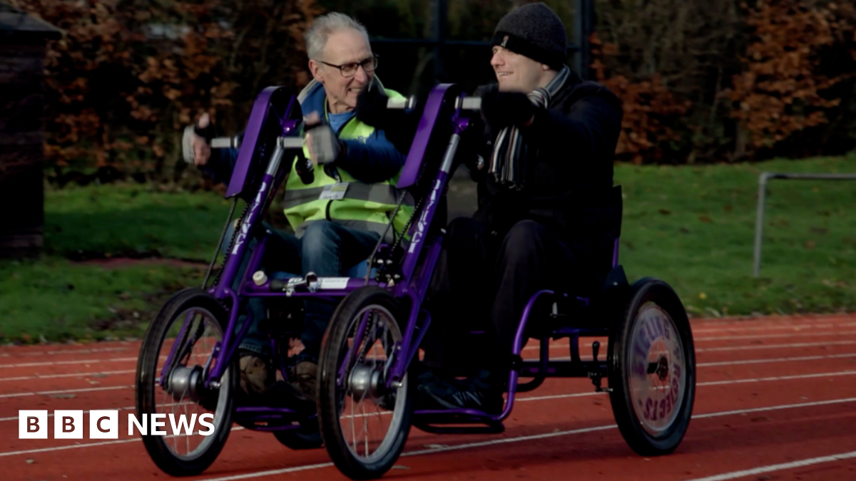 Two men, one on the left with grey hair and wearing glasses and another on the right wearing a hat, are sitting alongside each other on a type of bike, which is in part purple. It is on an athletics track and grass is in the background.