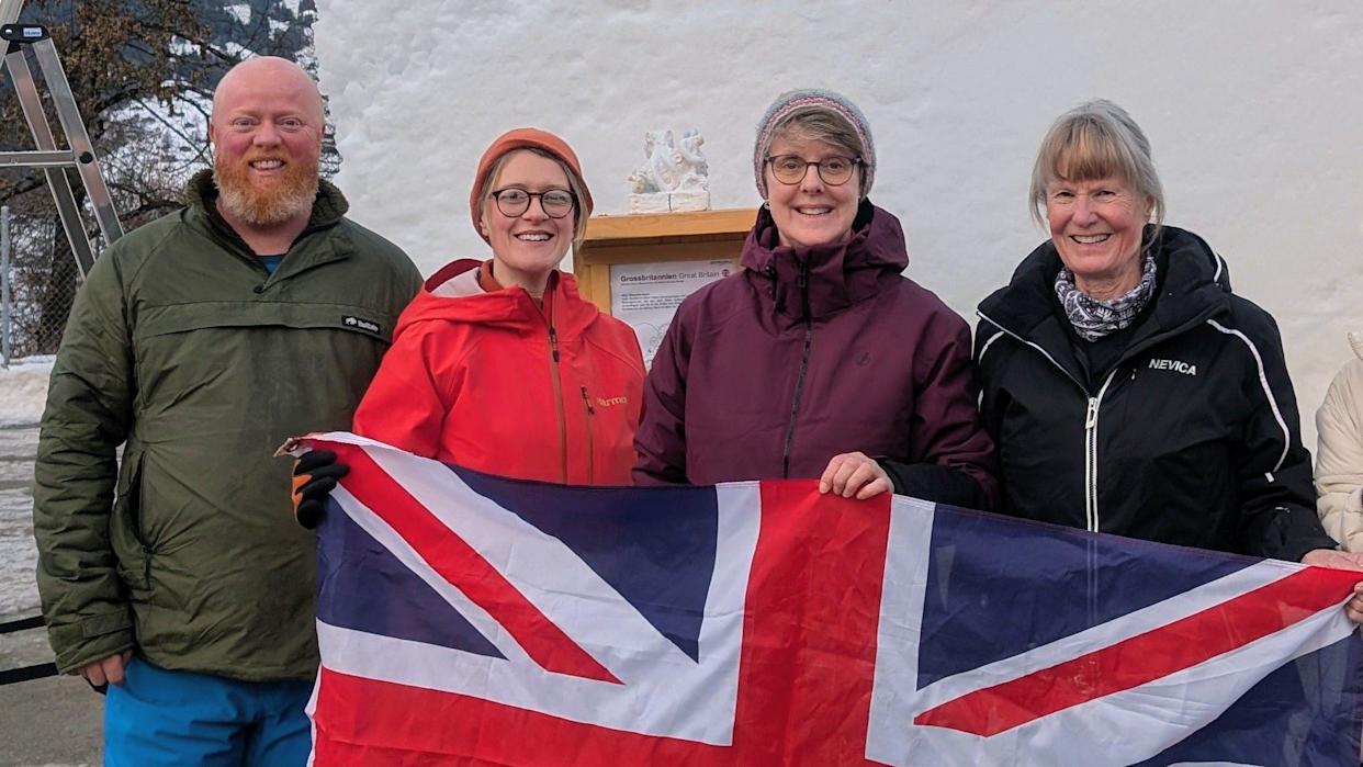 Three women and a man stand in front of a block of snow holding a Union Jack