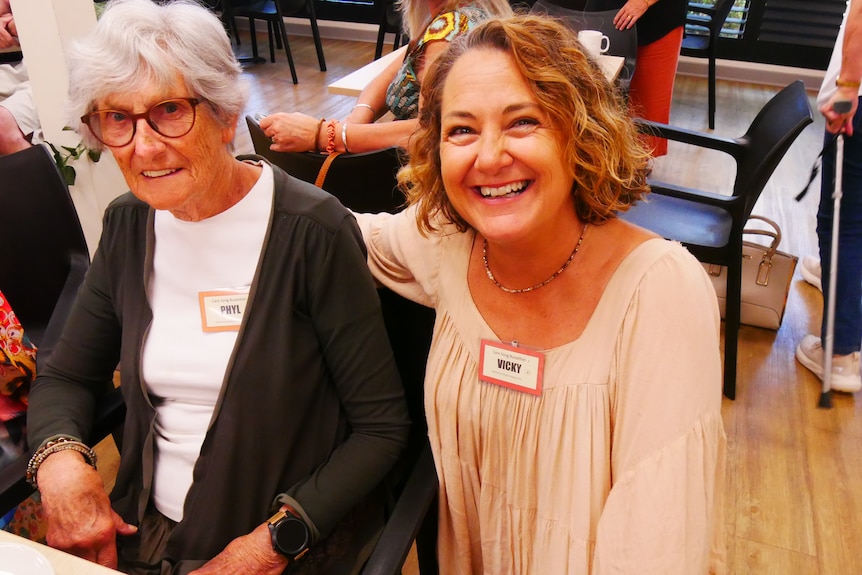 An older woman with short, white hair sits next to a smiling middle-aged woman in a common space.