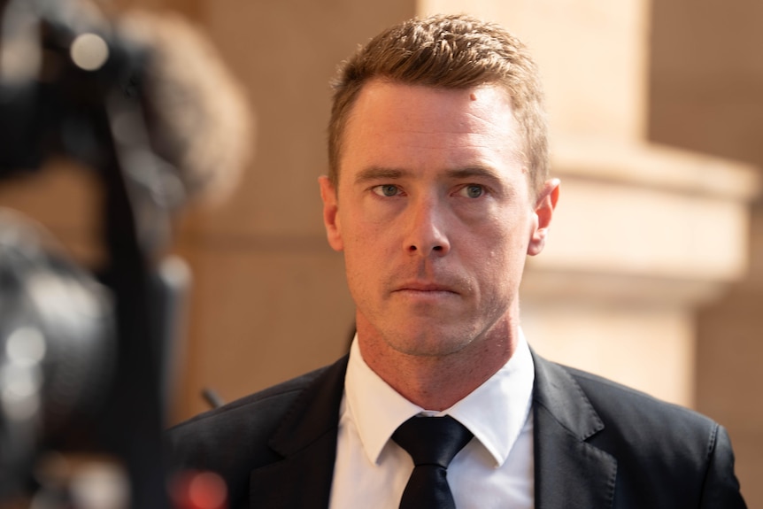 Rohan Dennis wearing formal attire walks along a sandstone wall, with a broadcast camera partially visible.
