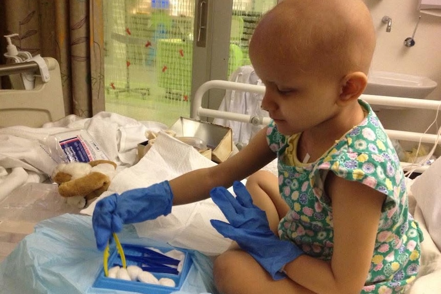 A young girl with no hair plays with medical supplies in a hospital bed.