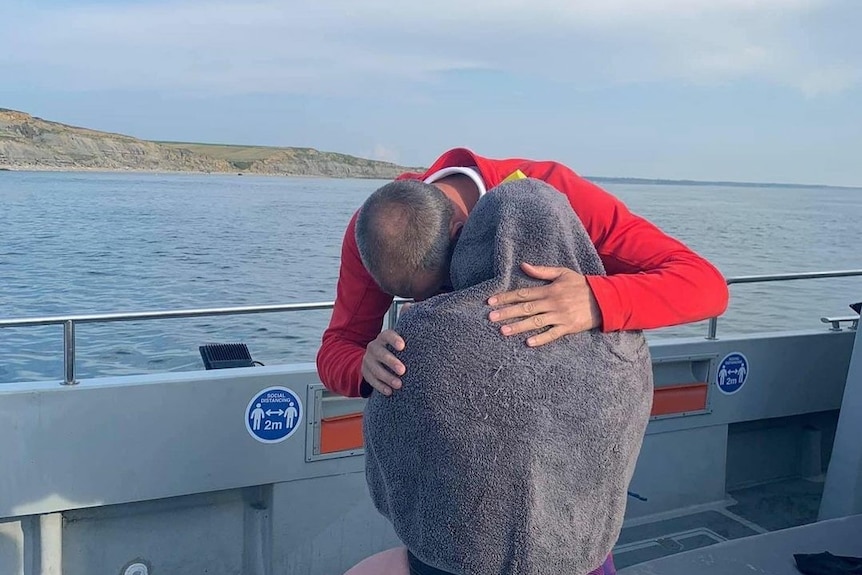 A woman sits on a boat wrapped in a grey towel, a man in a red jacket embraces her