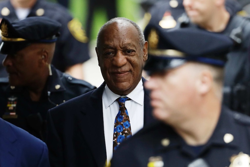 Bill Cosby, wearing a black suit and blue tie, stands in a crowd of police in navy blue uniforms. 