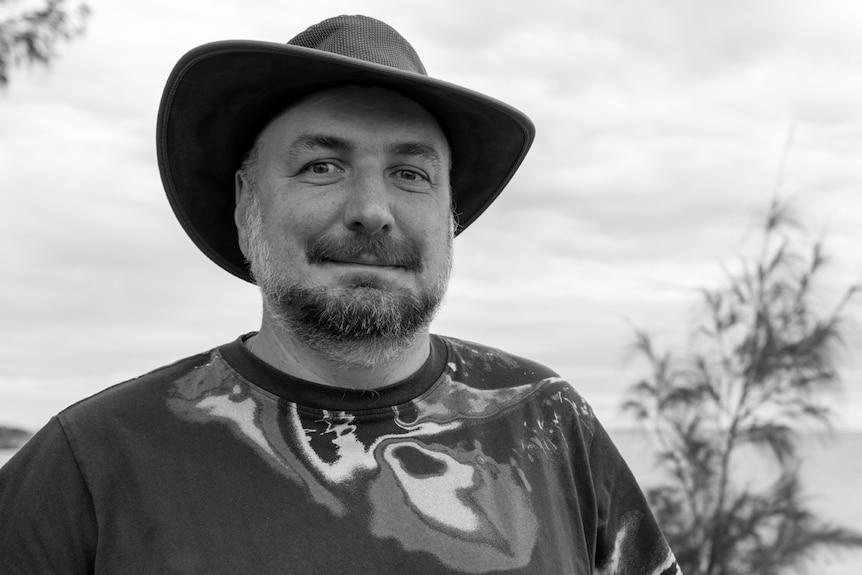 A black-and-white photo of a smiling man wearing a wide-brimmed hat and a T-shirt with the coastline behind him.
