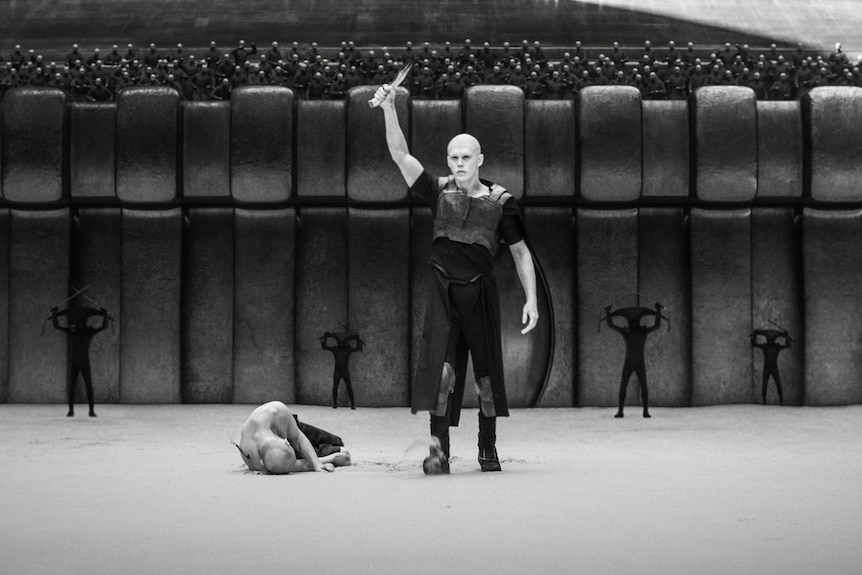 A man in an arena holds a knife above his head while his opponent lies on the ground