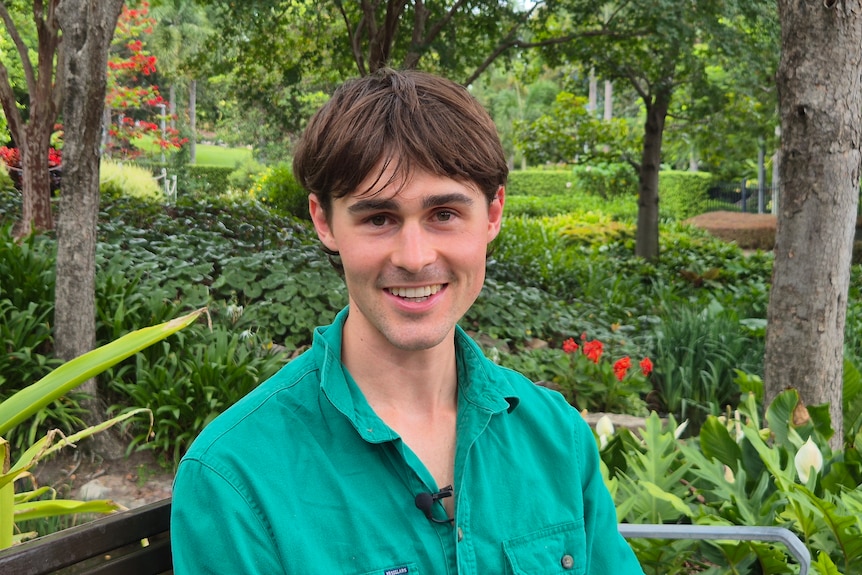 A man in a green shirt sits on a chair against a garden background
