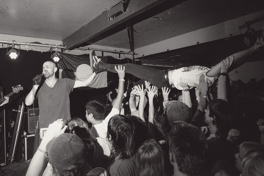 Black and white photo of musician on stage singing in front of lively audience with someone crowdsurfing over hands