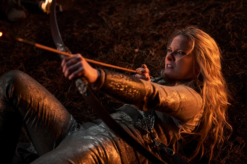 Still image of a woman in fantasy-style armour on her back on the ground, but determinedly holding an arrow to a bow