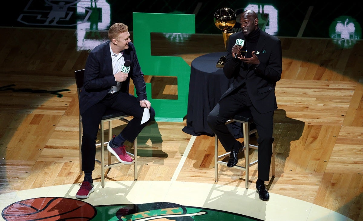 Two men are seated on stools on a basketball court, engaged in a conversation while holding microphones. One man wears a dark blazer, white shirt, black pants, and red sneakers, holding a piece of paper. The other man is dressed in an all-black suit with a turtleneck and black shoes. Behind them is a large green number 5 and a table with a basketball championship trophy. The setting suggests a formal interview or discussion event related to basketball.