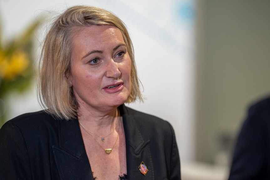 A woman with short blonde hair wearing a black blazer looks off camera, speaking