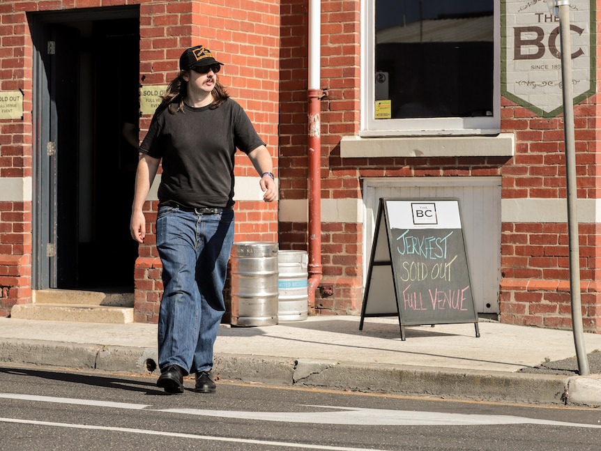 Man with sunglasses walks past a chalk sign outside a pub on a street corner that reads Jerkfest Sold Out Venue