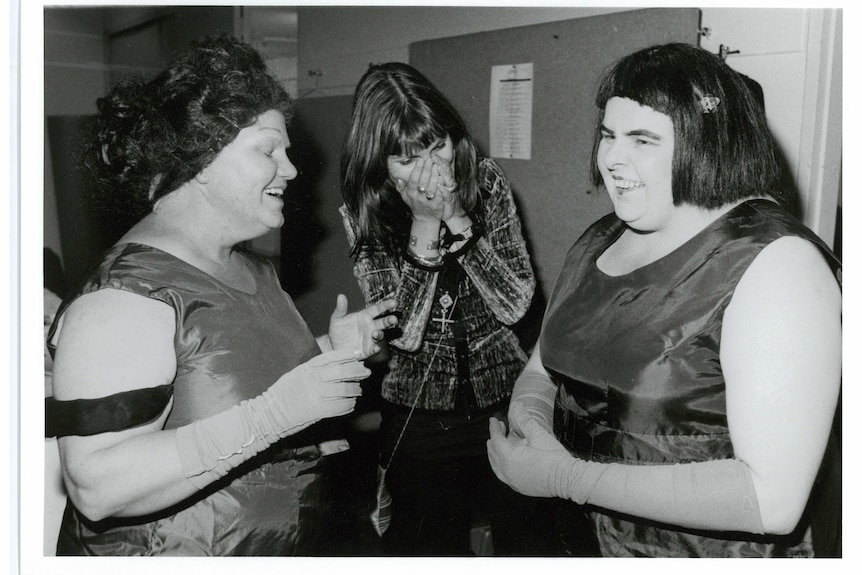 Two women dressed in wigs, dresses and gloves smile facing each other while a third woman laughs in the background.