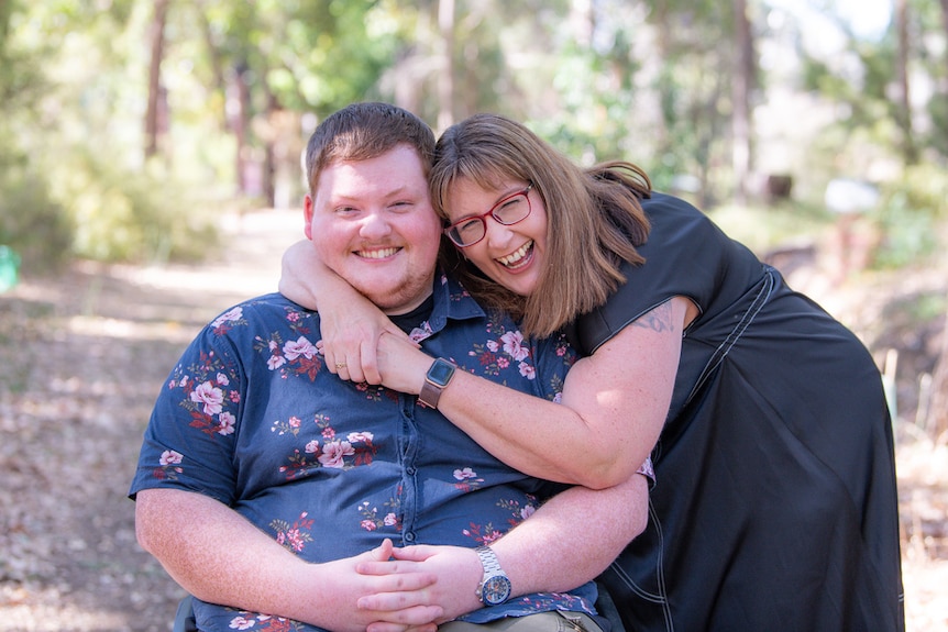 A mother and sun embrace and smile at the camera. They are outside in the sunshine.