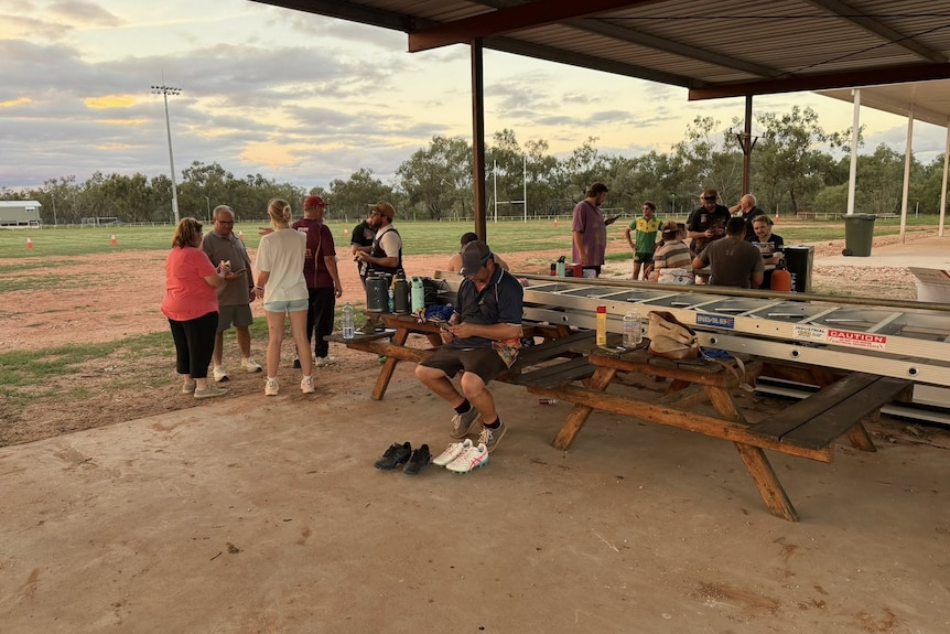 People sitting and standing around picnic table at footy field. 