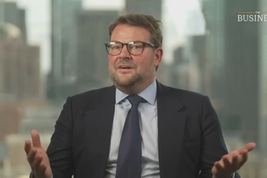 Charlie, is Caucasian, wears glasses, dark suit and blue tie, light shirt, has slight beard, gestures with hands.